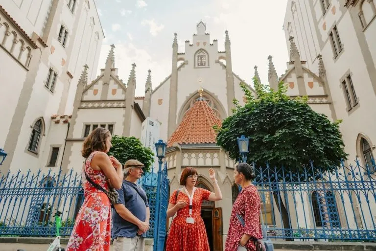 a group of tourists standing in front of the Maisel Synagogue in Prague listening to their local tour guide pointing at the intricately decorated roof of the synagogue