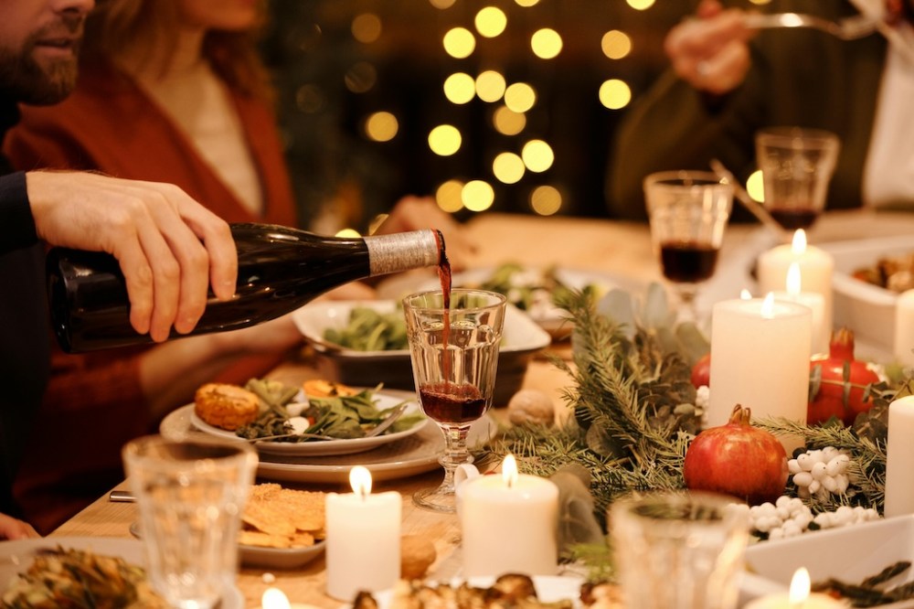 a person sitting at a table decorated for Christmas with another person pouring red wine into the prepared wine glasses