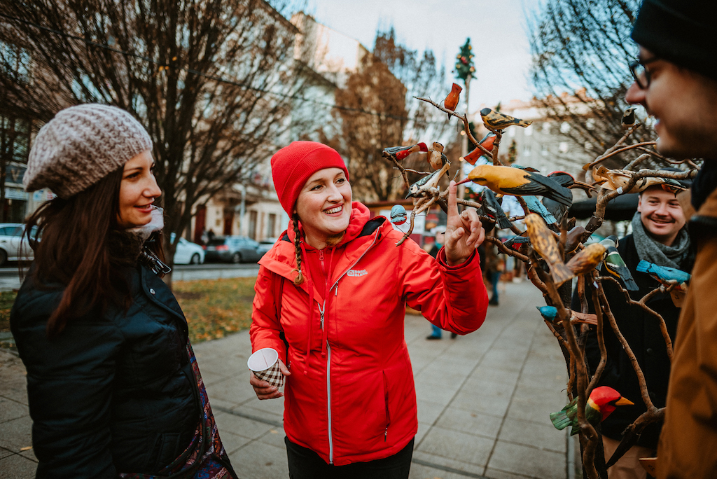 A Prague City Adventures tour guide in a red winter jacket pointing at hand-made wooden birds to a group of tourists at one of the stands on Prague's Christmas Markets